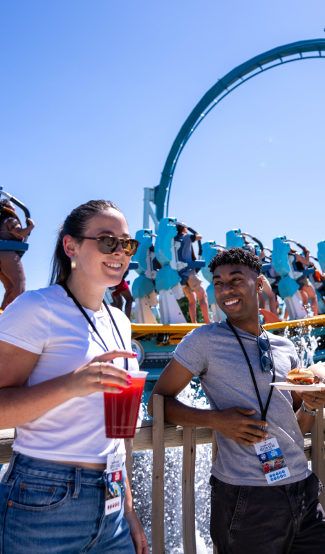 Two people with food and drinks in front of a roller coaster