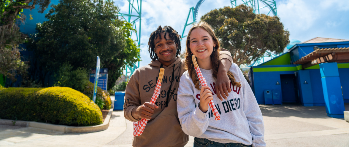 Two teens holding churros in front of a roller coaster